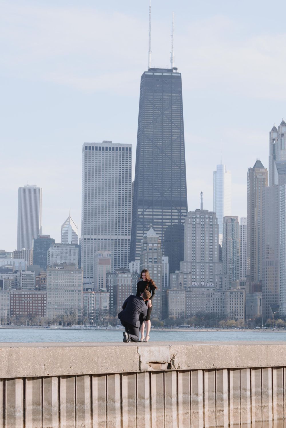 Someone sits perched on a concrete wall along Lake Michigan with Chicago's iconic skyline and John Hancock building in view.