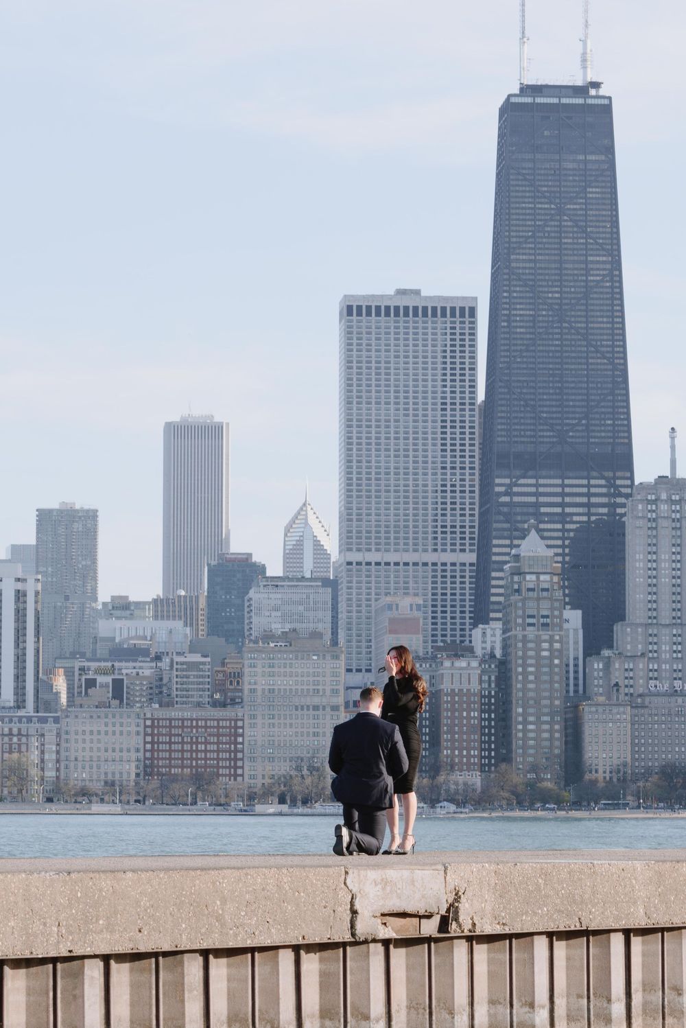 Two figures stand on a concrete pier overlooking Lake Michigan and Chicago's iconic skyline featuring the John Hancock Center.