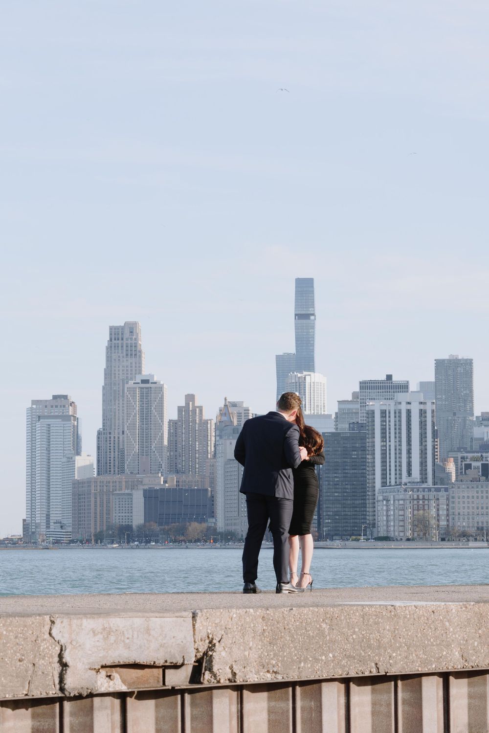 A couple stands on a concrete ledge overlooking the Manhattan skyline and East River on a hazy day.