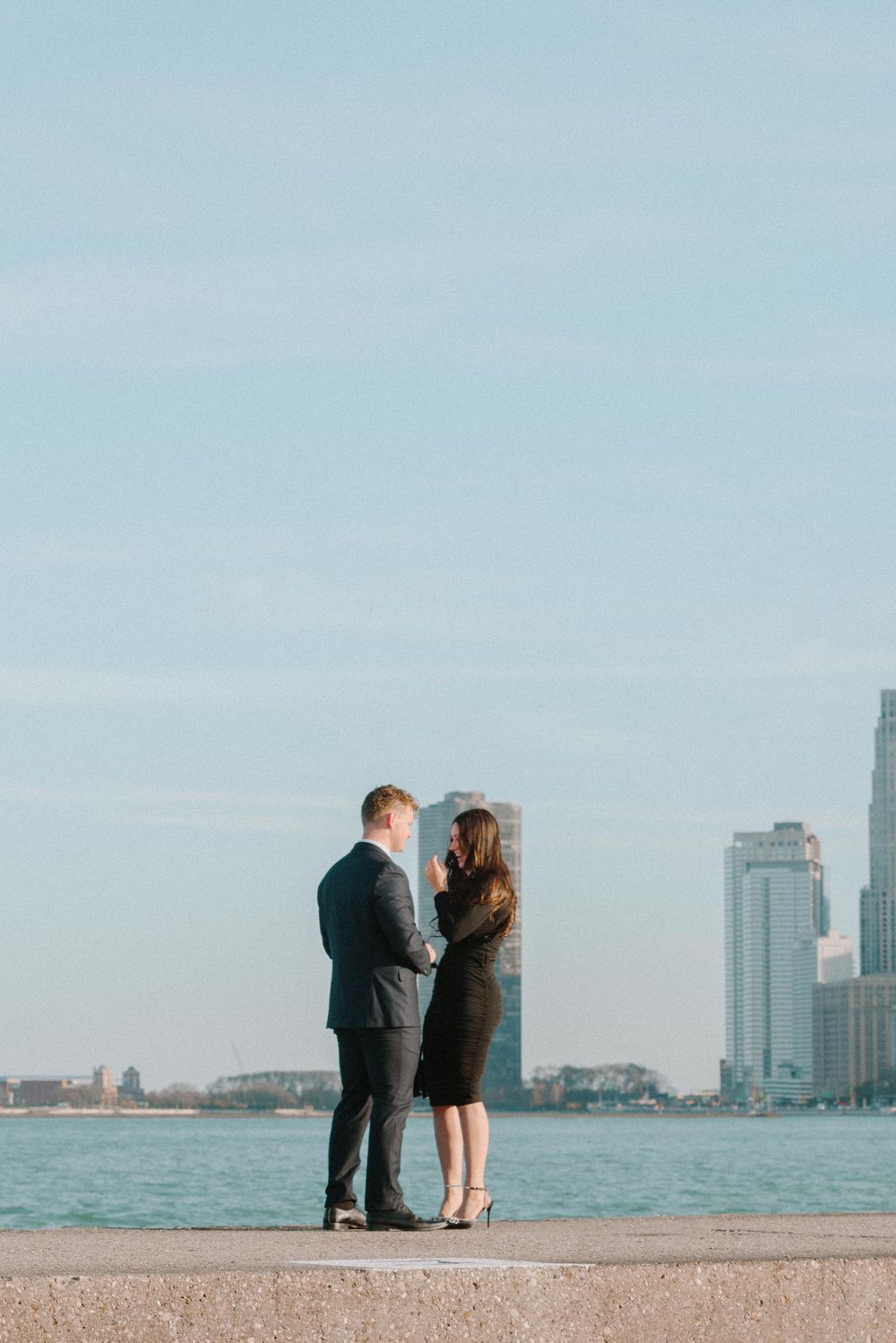 A couple embraces while standing on a concrete pier overlooking Lake Michigan with the Chicago skyline in the background.