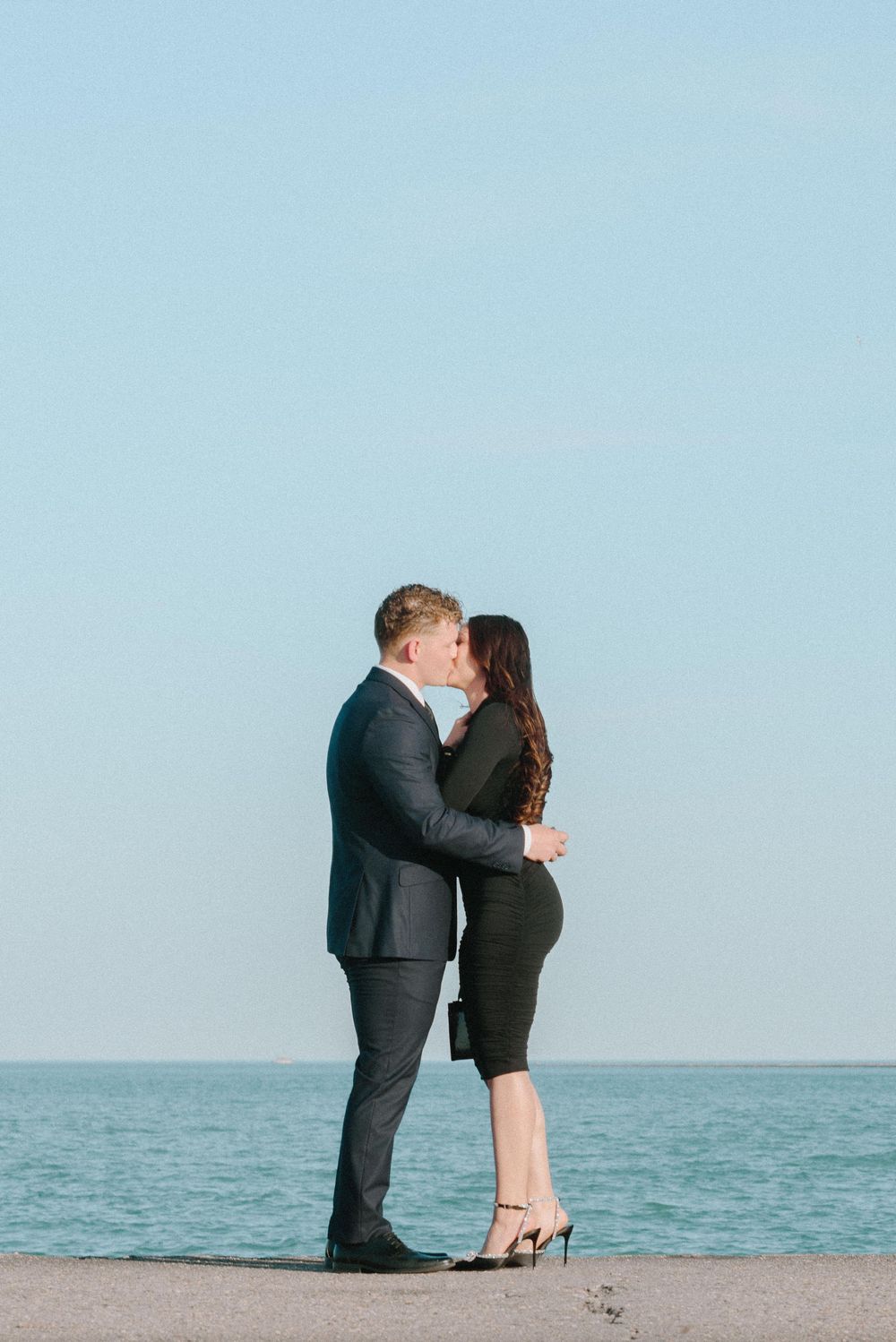 A romantic couple embraces and kisses by the ocean on a serene day with blue skies.