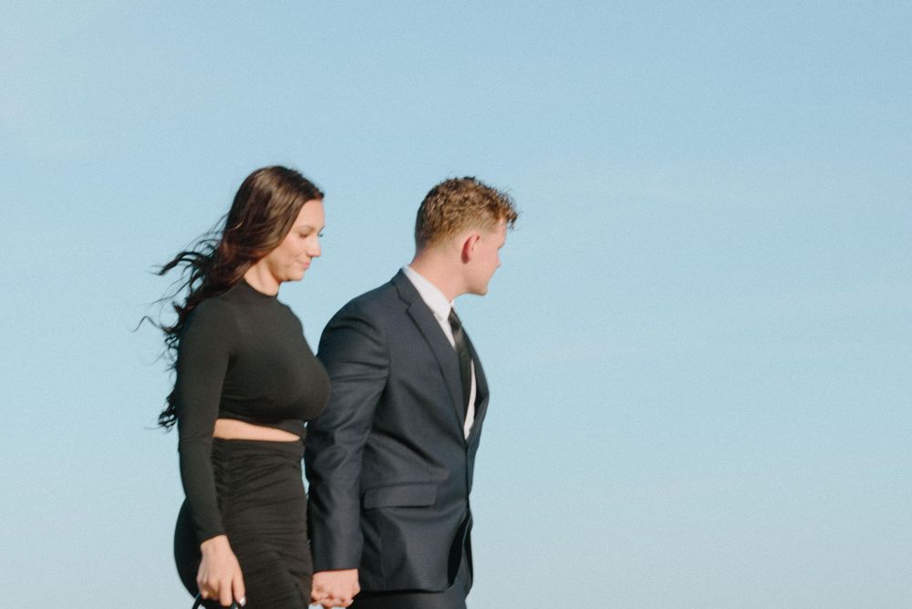 A couple in black formal attire holding hands while standing against a bright blue sky.