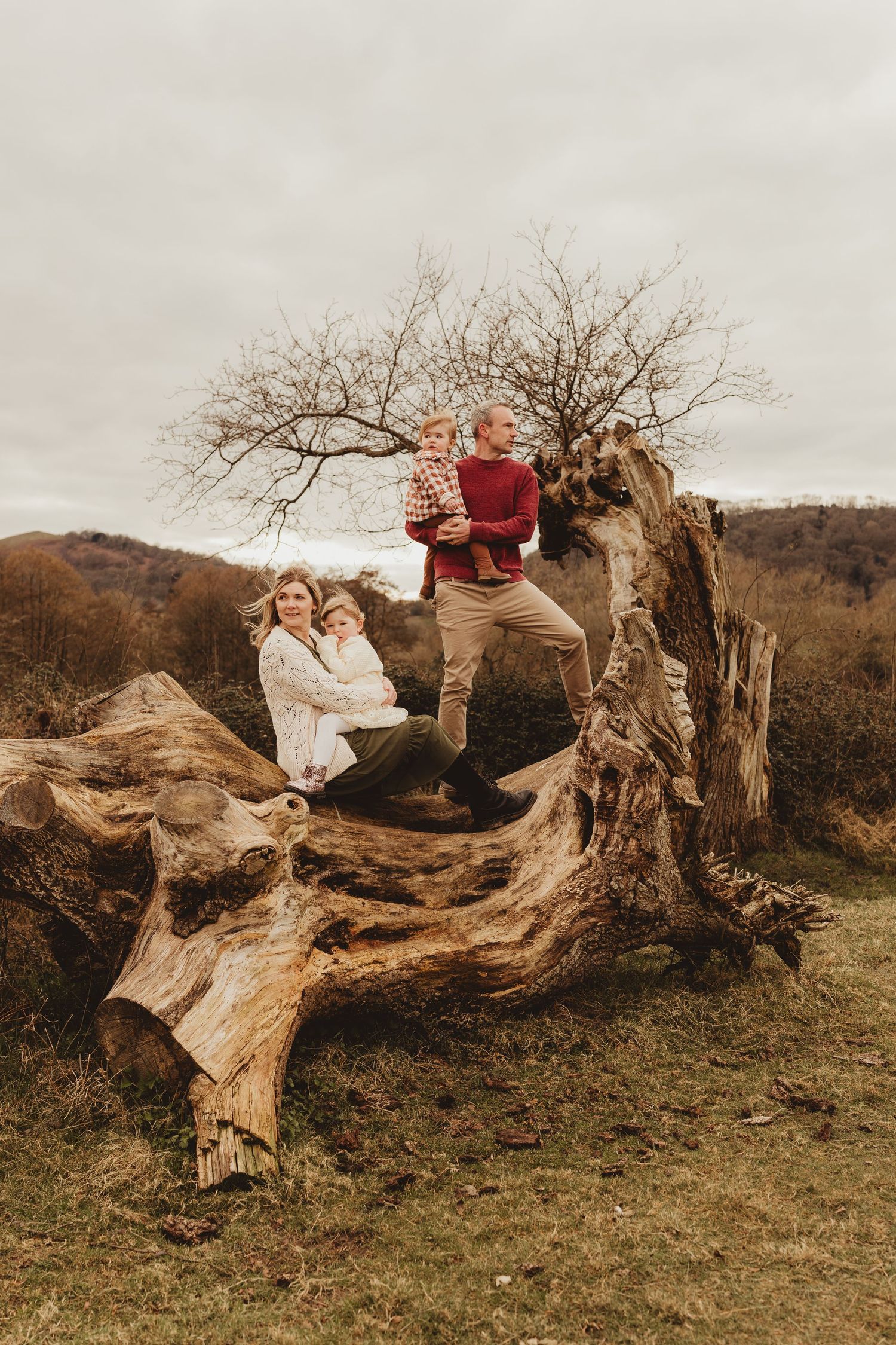 A group sits together on a large fallen tree trunk against an autumn mountainous landscape on an overcast day.