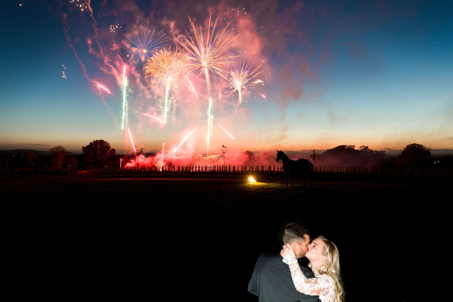 Romantic couple embraces while watching vibrant fireworks display lighting up the twilight sky over dark landscape.