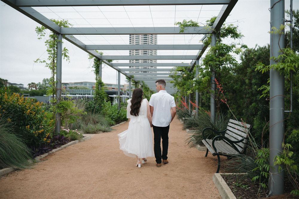 Couple walks hand in hand down a romantic garden path under a modern metal pergola covered with greenery.