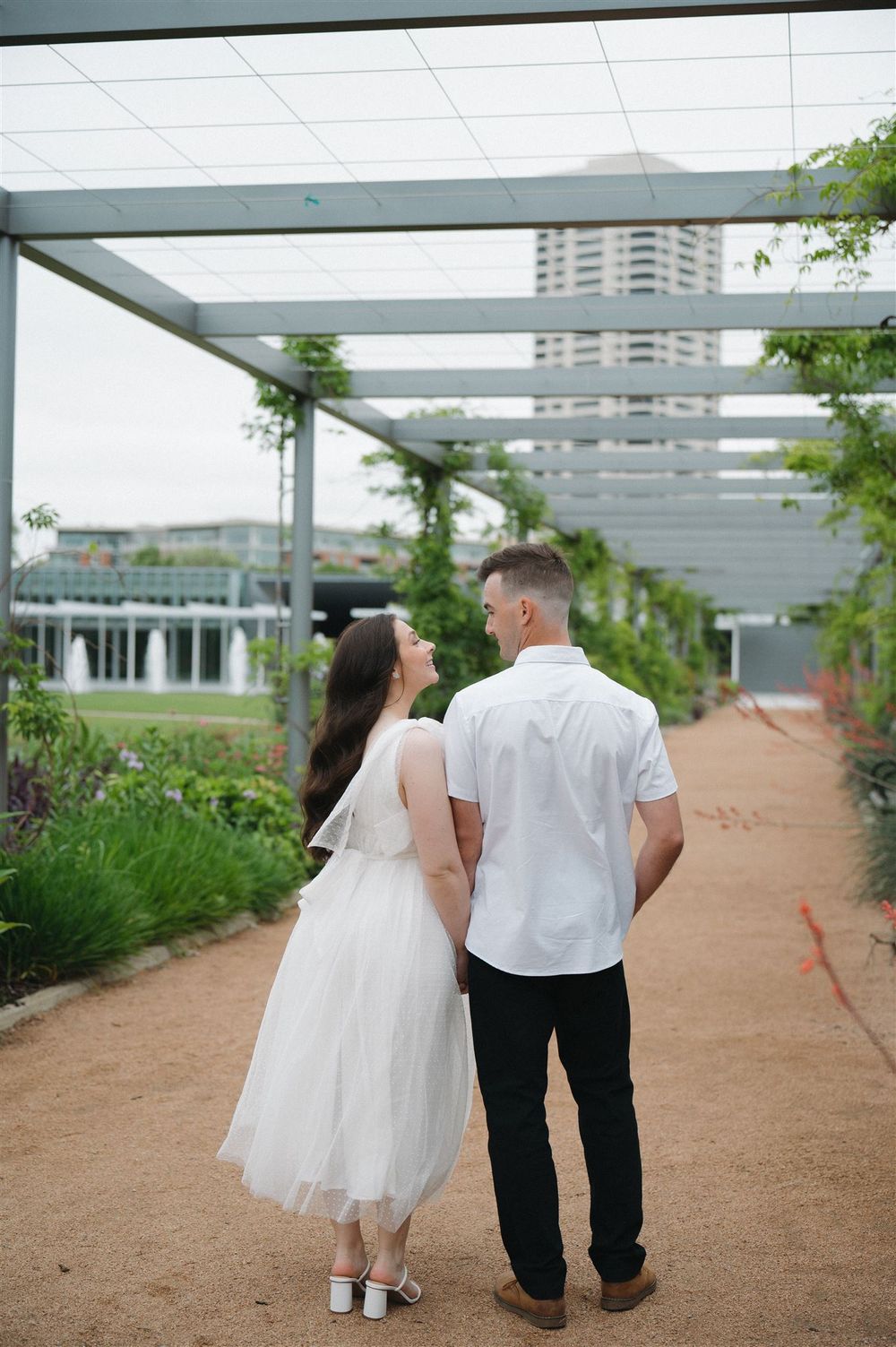 A couple walks down a garden path beneath a modern pergola structure with a city building visible in the background.