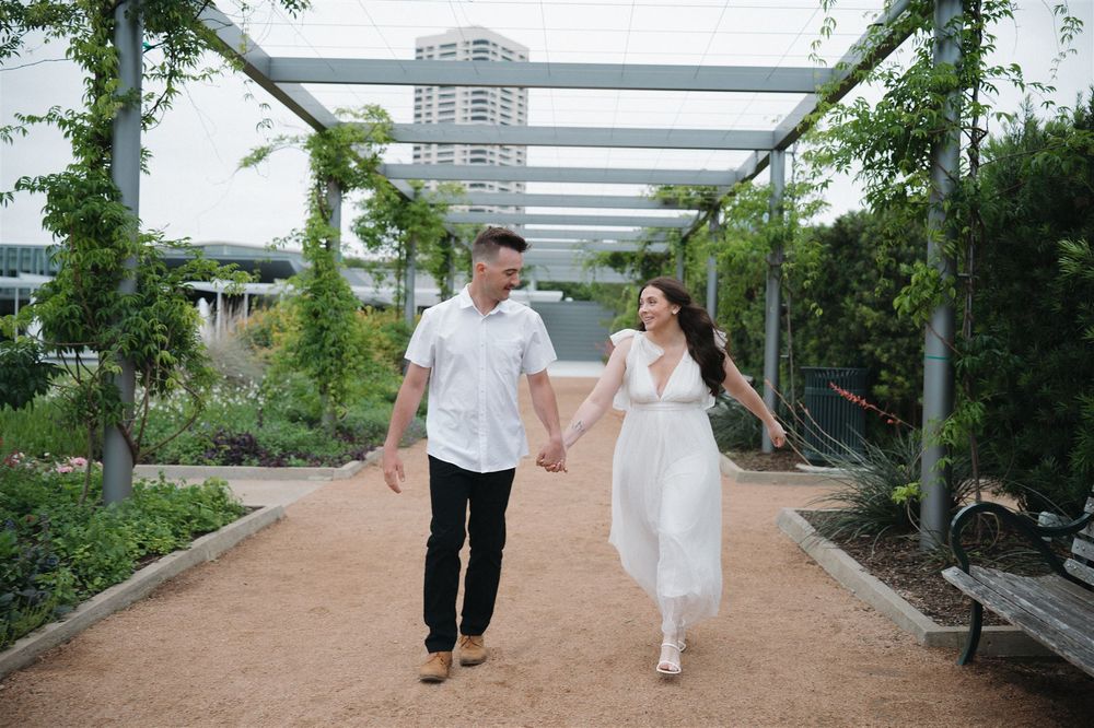 A couple walks hand in hand along a garden path beneath a modern pergola covered in climbing vines.