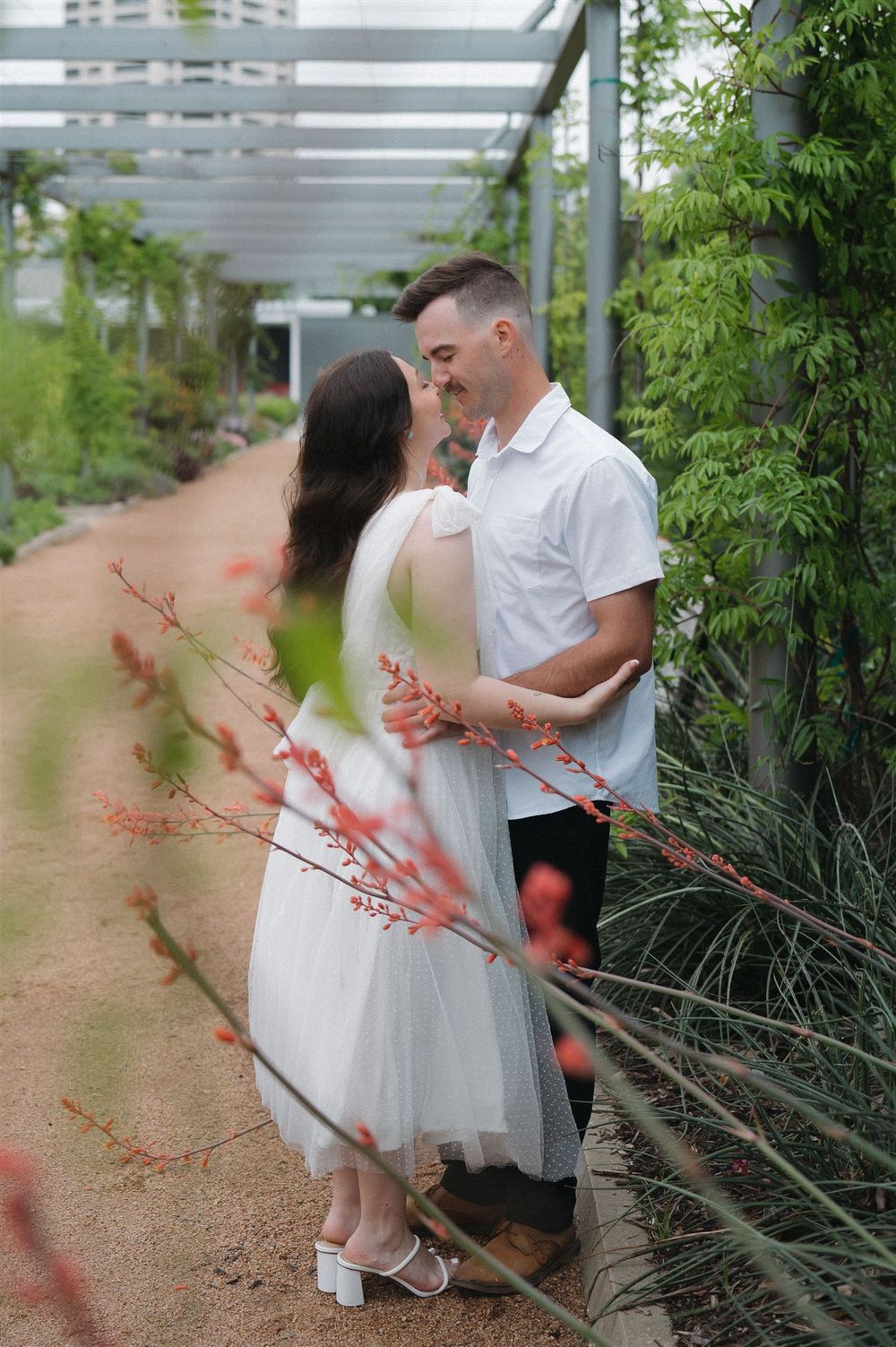 A couple shares an intimate moment in a garden walkway with white pergola and greenery surrounding them.