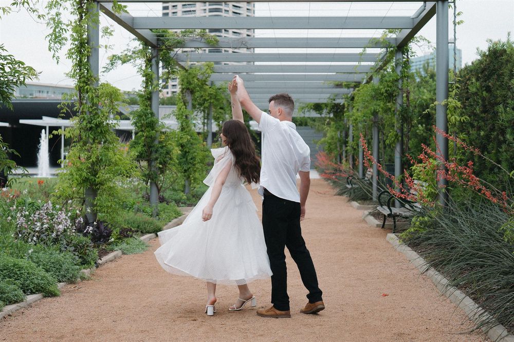 A couple walks along a garden path under a green pergola covered in climbing vines and flowers.