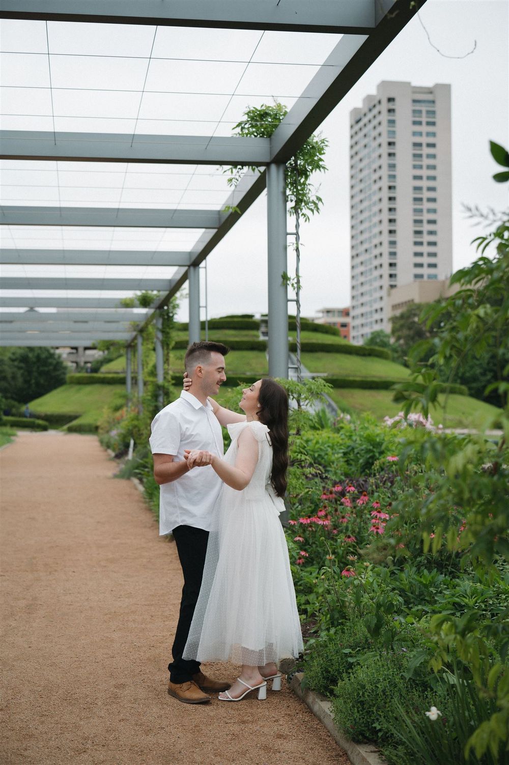 A couple shares a romantic moment under a pergola walkway surrounded by gardens with a high-rise building in the background.