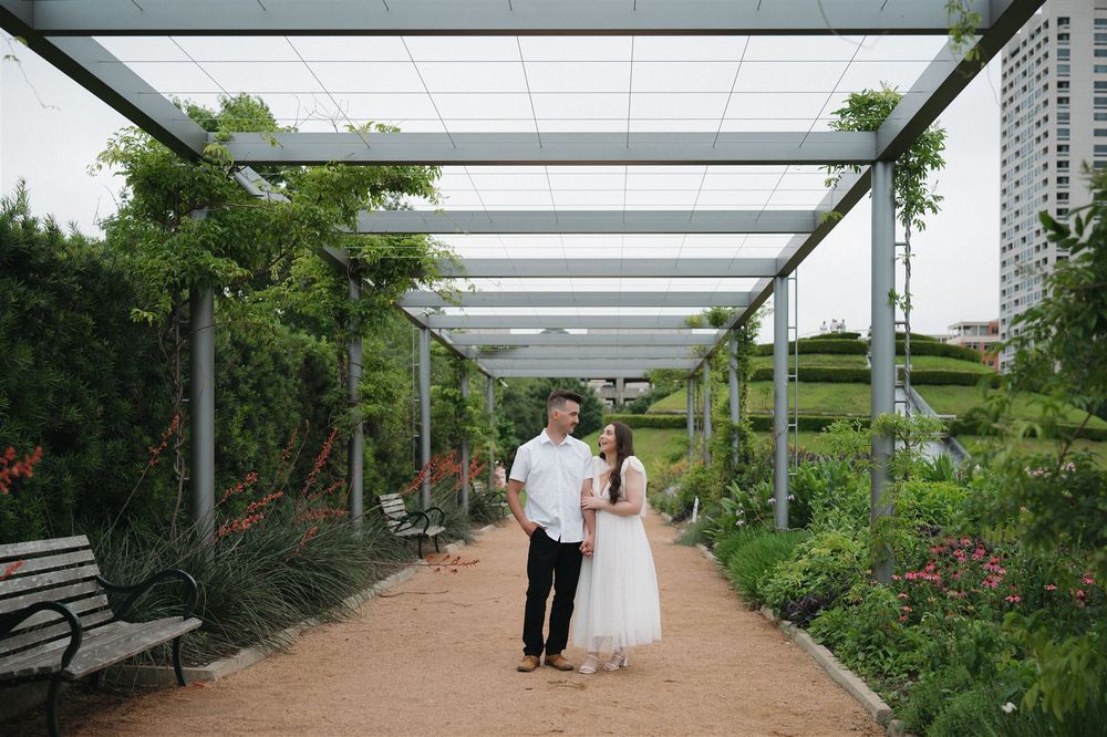 A couple walks hand in hand along a covered garden walkway with benches and lush greenery on a sunny day.