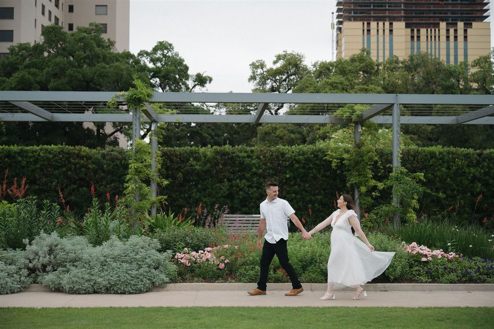A couple walks hand in hand through a lush garden setting with greenery and architectural elements in the background.