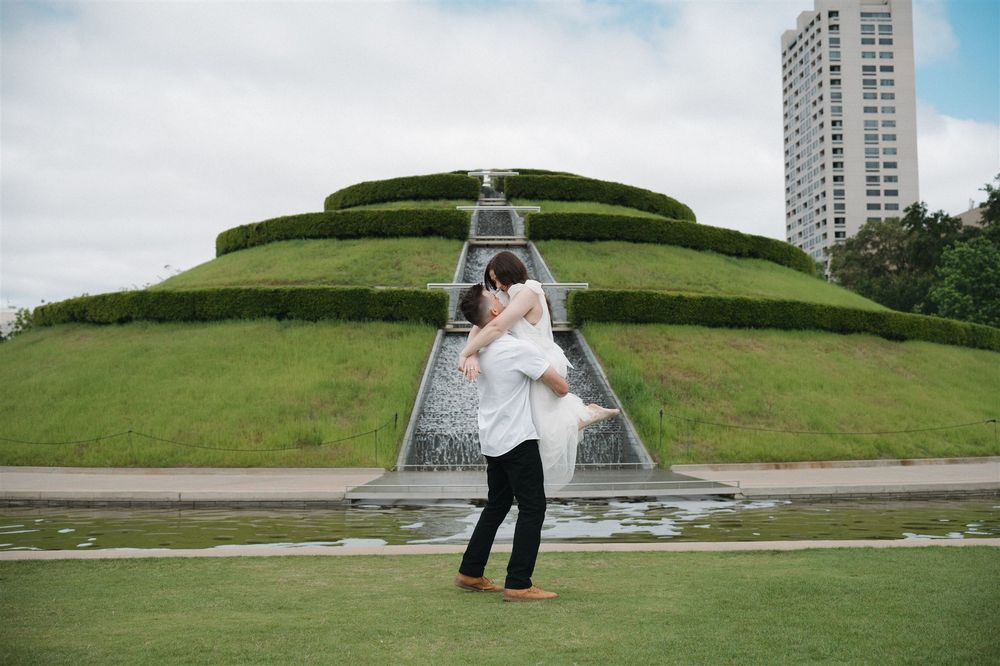 A couple embraces on a pathway leading to a tiered green hill structure with a modern high-rise building in the background.
