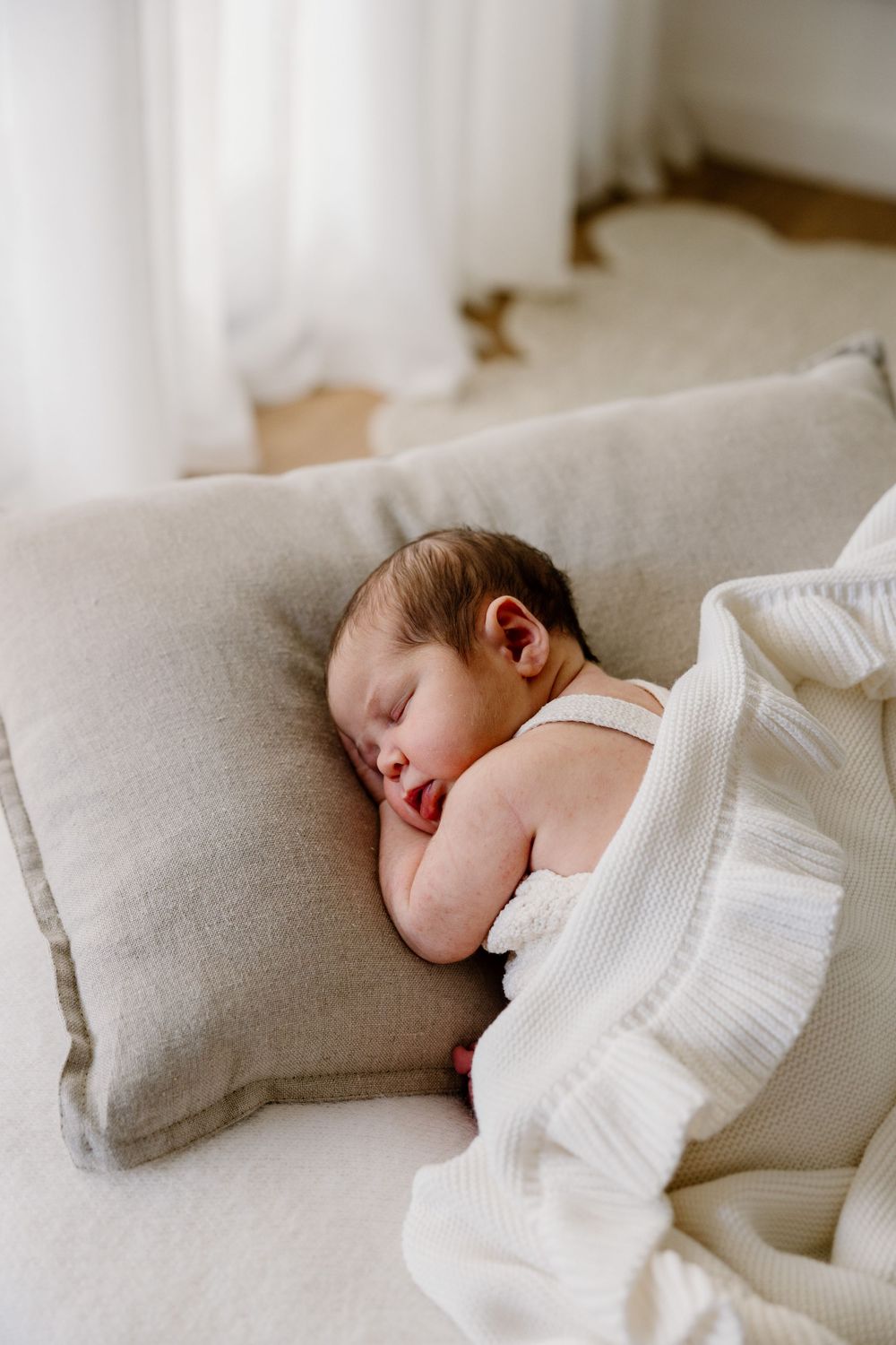 Peaceful sleeping baby wrapped in white blanket resting on gray cushion in soft natural light.