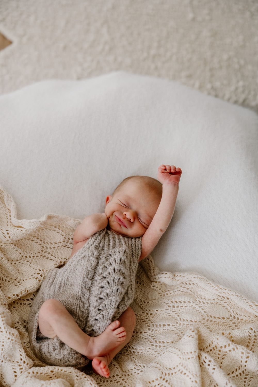 Sleeping newborn baby stretches on white bedding with knitted beige blanket in soft natural light.