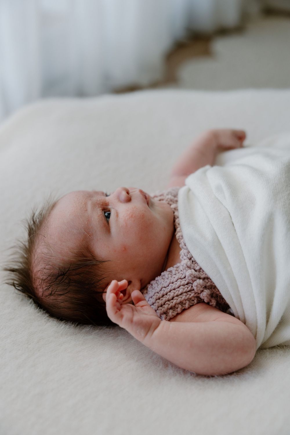 Baby with dark hair lying on white bedding wearing a knitted pink sweater looks to the side with peaceful expression.