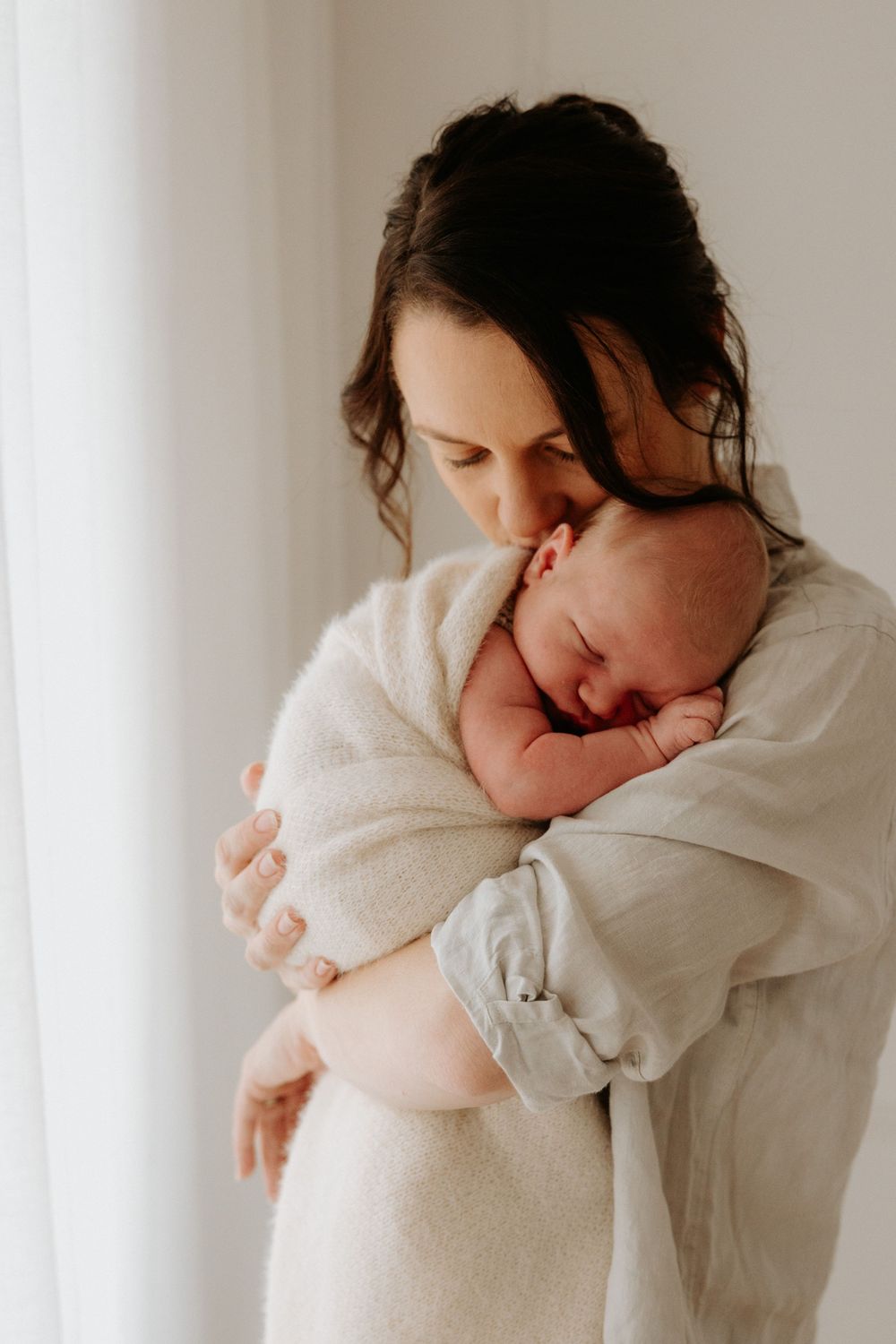 Tender and intimate image showing a baby swaddled in white being held in soft window light.