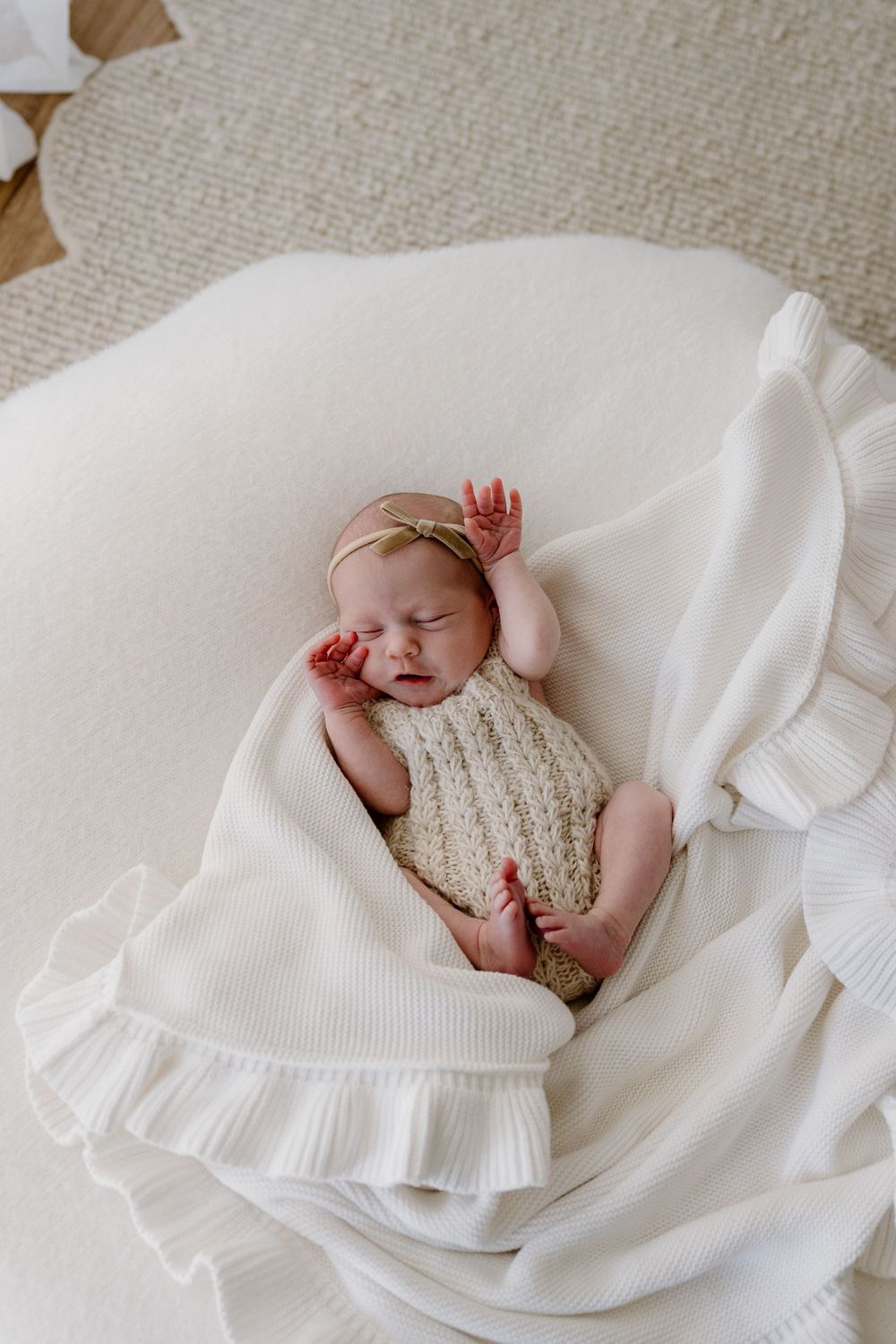 Newborn baby sleeping peacefully on white ruffled blanket wearing knitted romper.