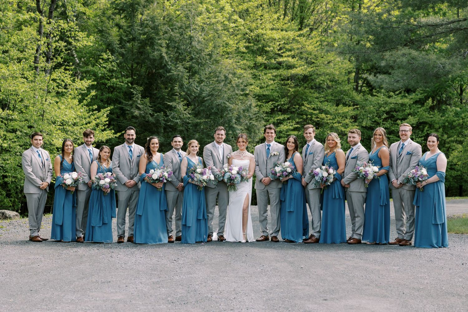 Wedding party in matching grey and blue attire poses together on outdoor path surrounded by lush green trees at Windham Manor.