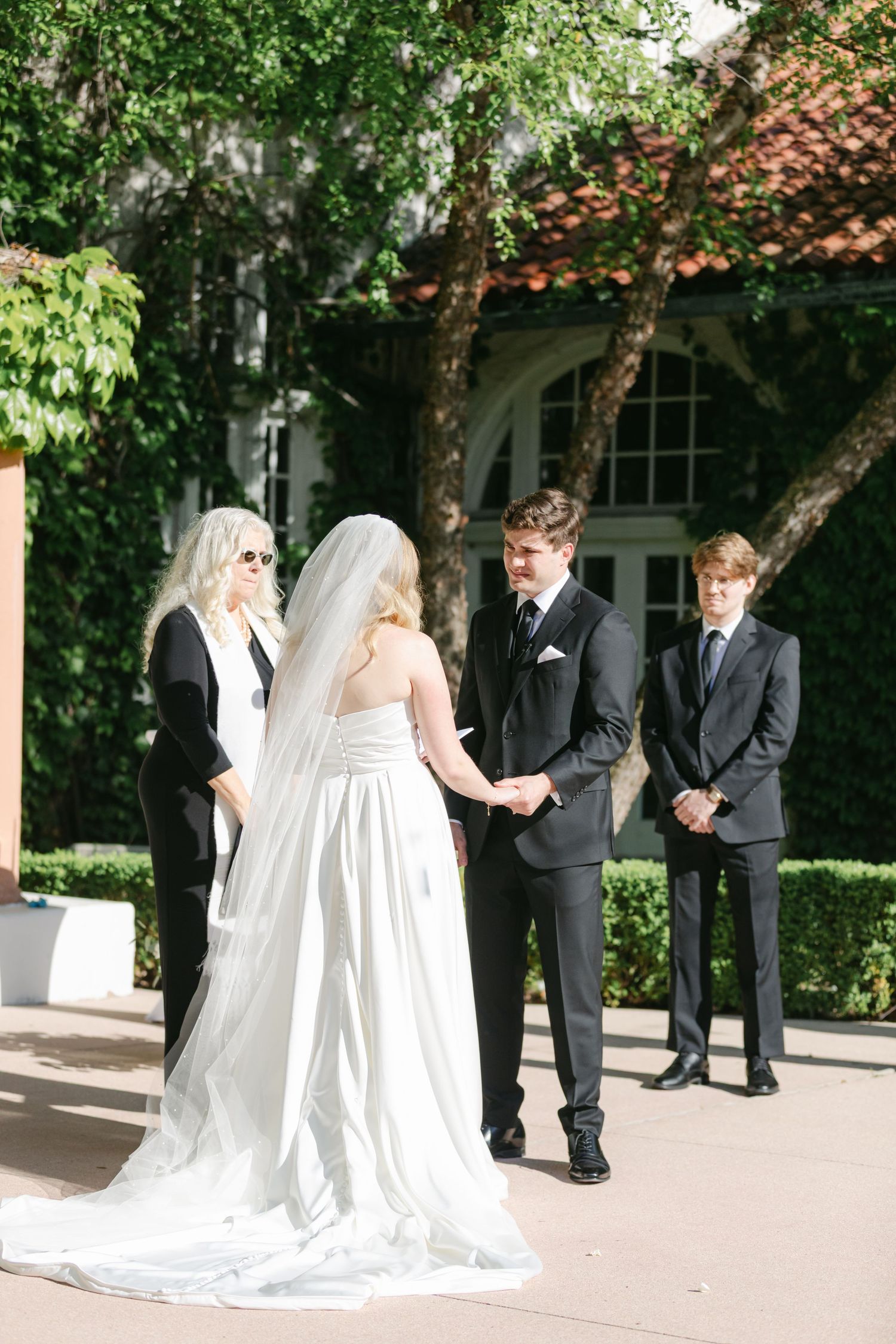An outdoor wedding ceremony with attendants in black dresses and the couple at the altar under trees with terracotta roofing.