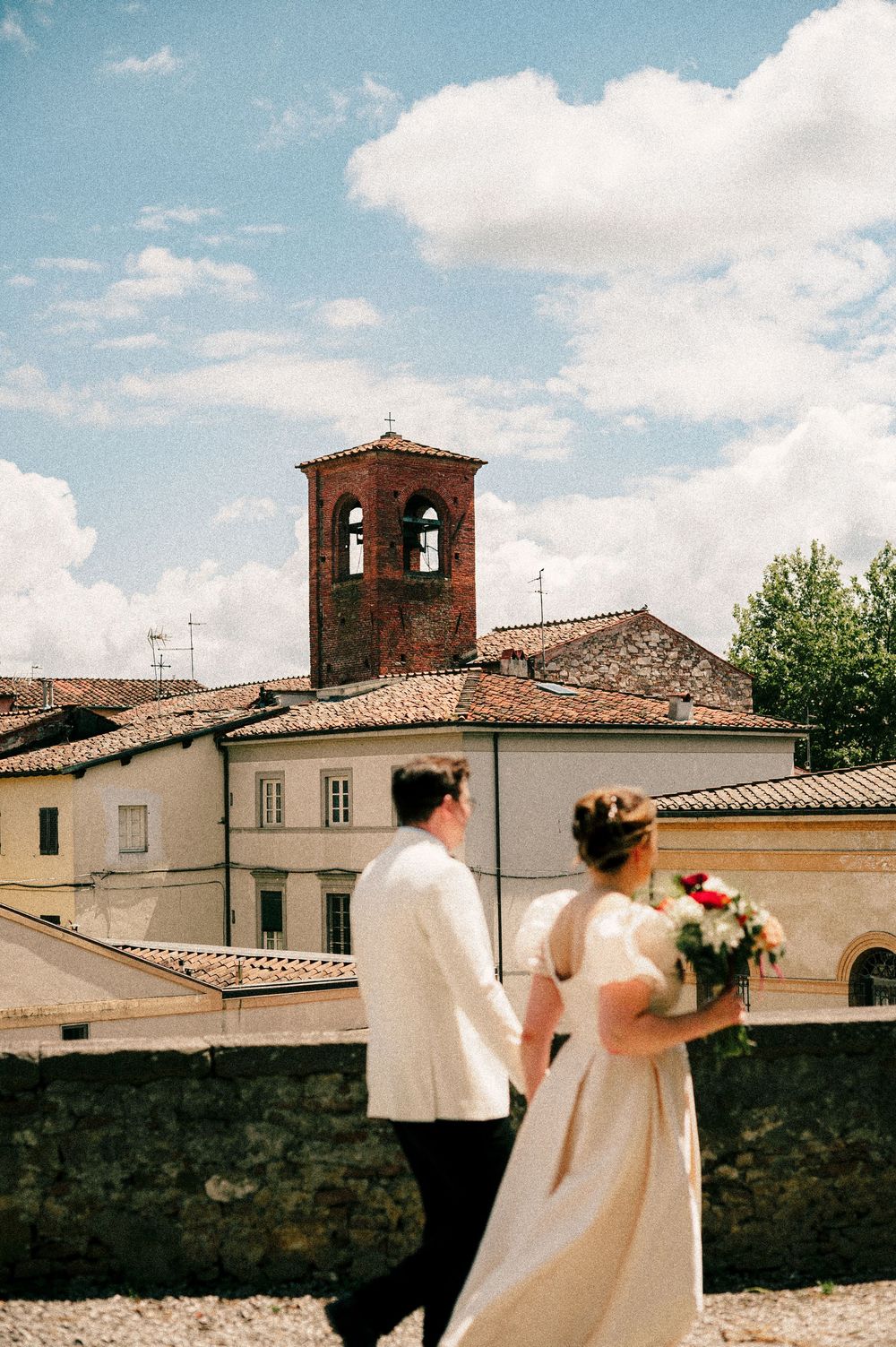 A couple in wedding attire views historic architecture with bell tower against blue sky.