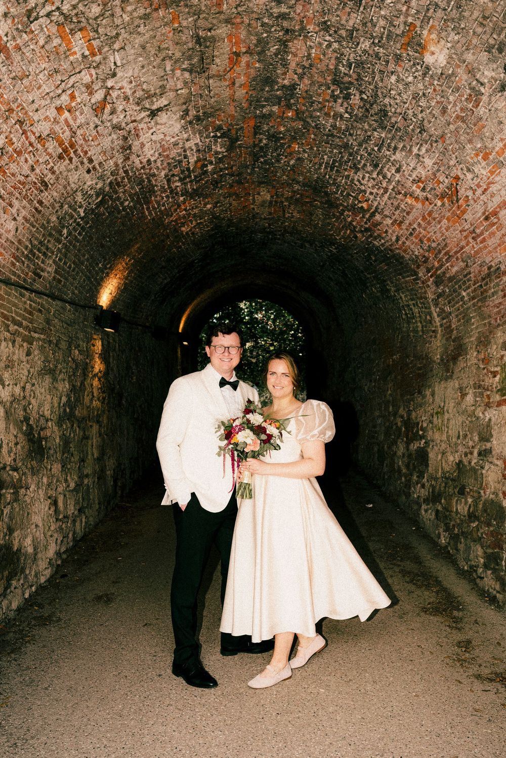 A couple poses for wedding photos inside a brick tunnel with natural light at the end.