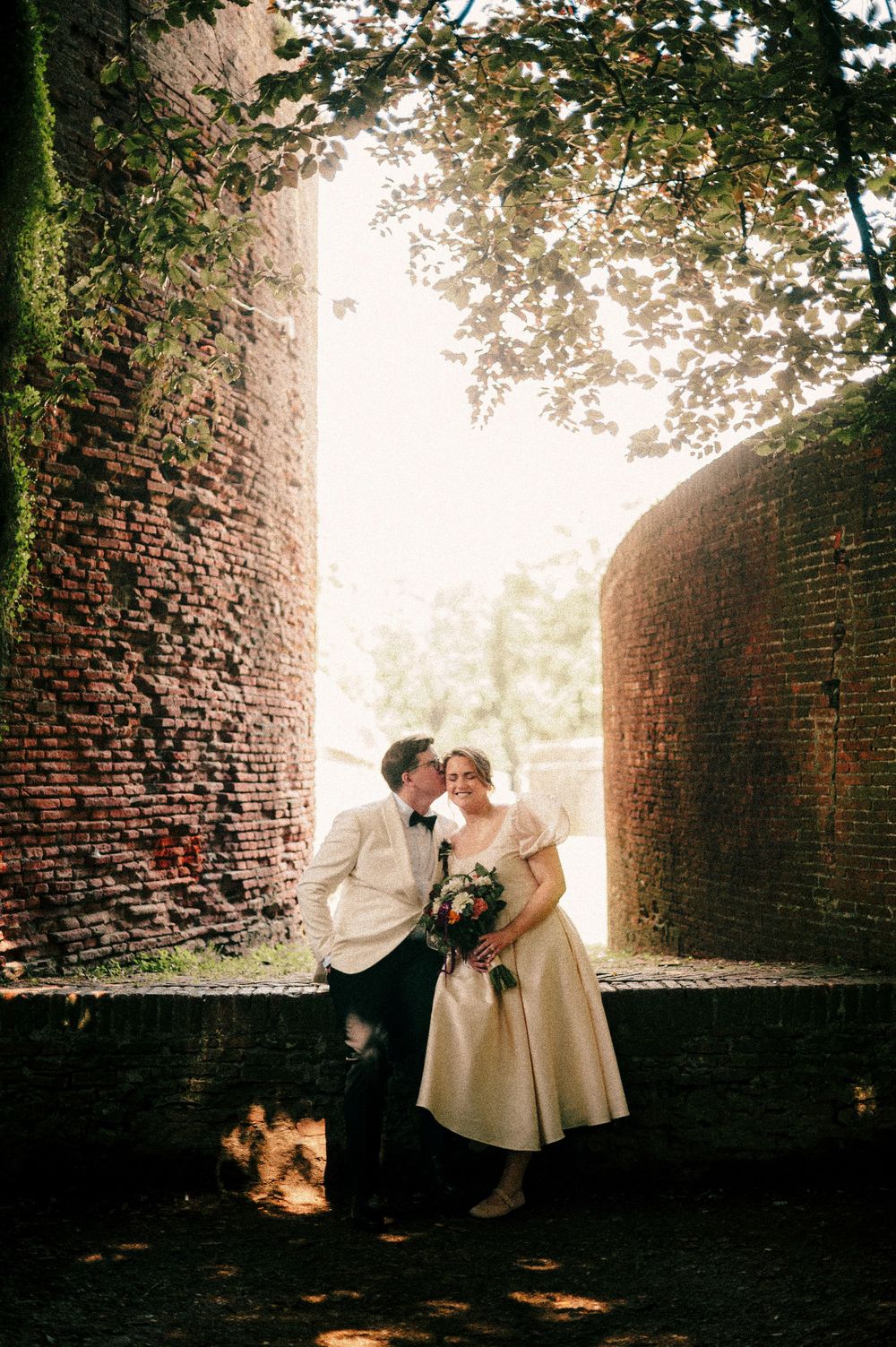A wedding photo between ancient brick walls with sunlight filtering through trees.