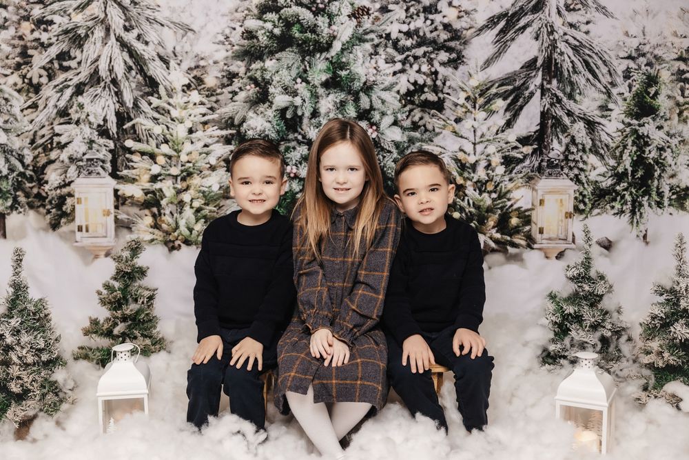 Three children wearing dark winter outfits pose on white chairs against a snowy winter forest backdrop with holiday decor.
