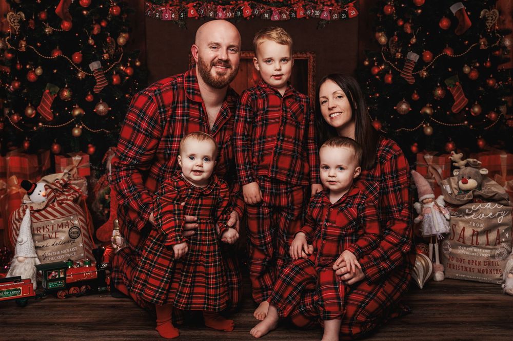 Family wearing matching red and black buffalo plaid flannel outfits pose in front of Christmas decorations.