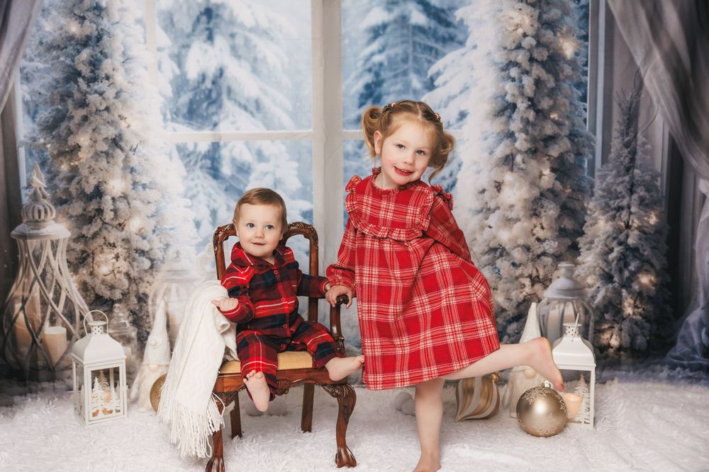 Children in red plaid outfits pose in a wintry Christmas scene with snow-covered trees and holiday decorations.
