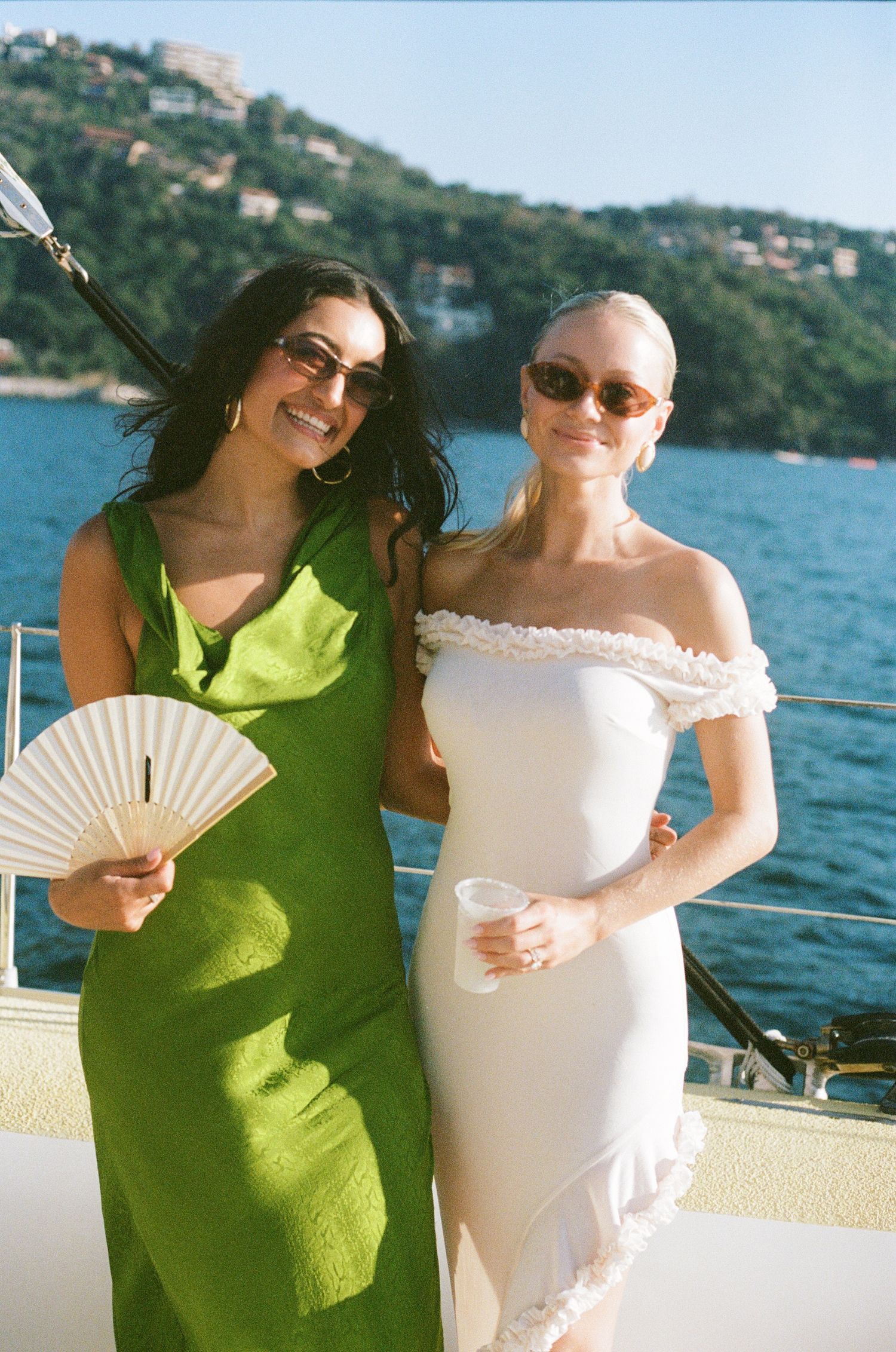 Two guests in formal dresses pose on a yacht deck with coastal scenery in the background.