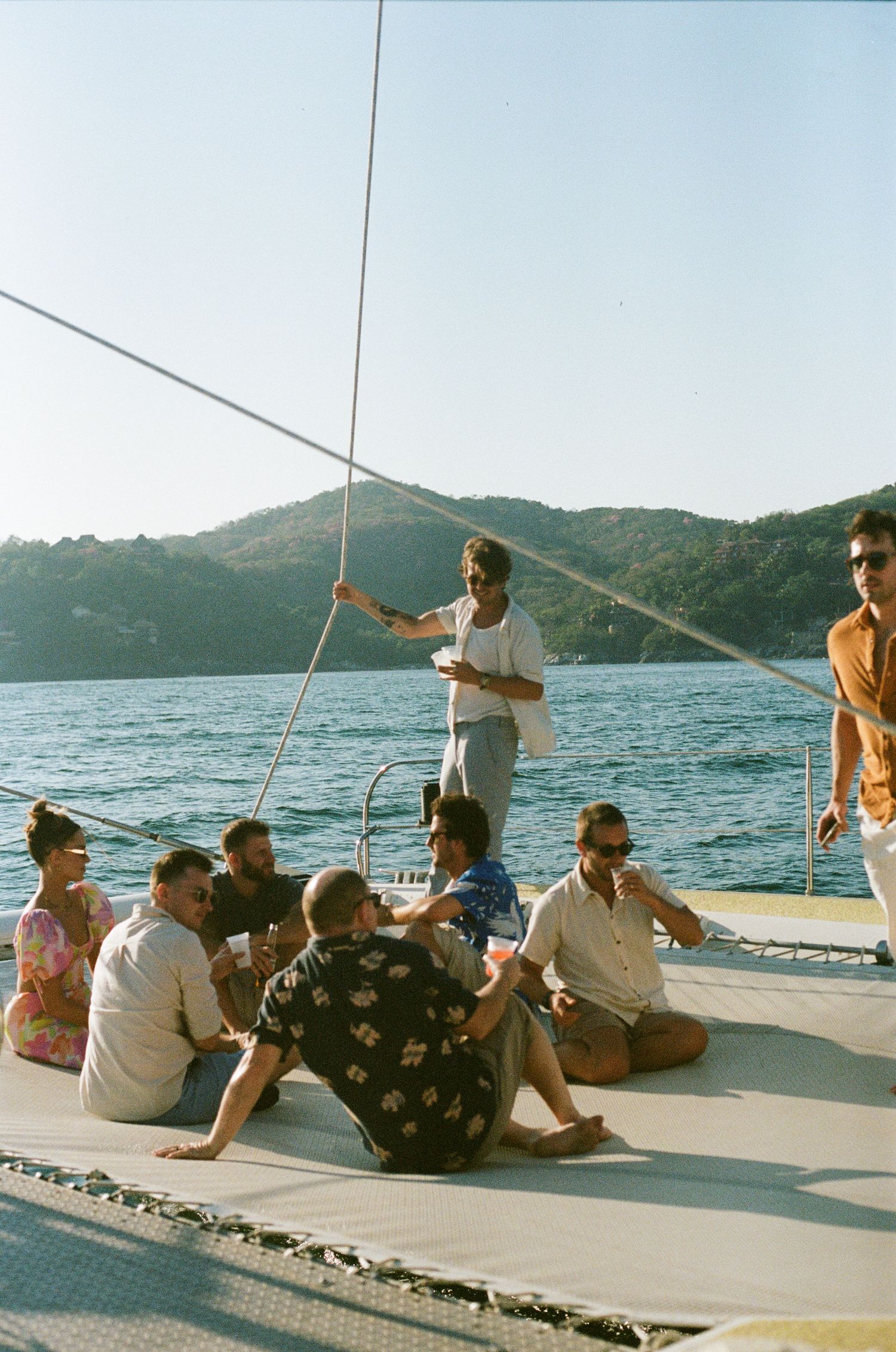 Group enjoying a sunny day on a catamaran deck with mountainous coastline views.