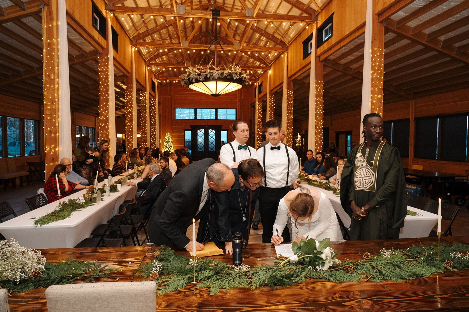 Wedding celebration in rustic wooden barn venue with guests gathered around long communal table with evergreen garland decor.