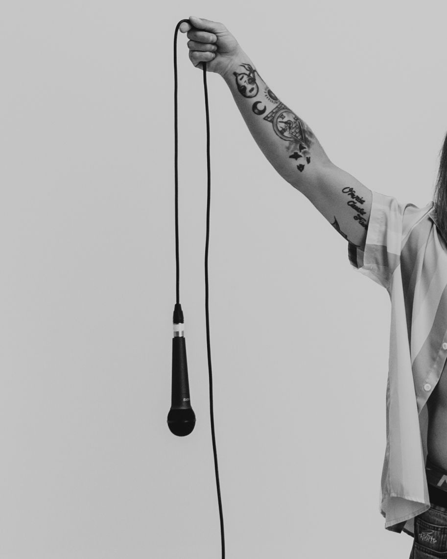A tattooed arm holds up a black microphone on a cord against a minimalist white background.