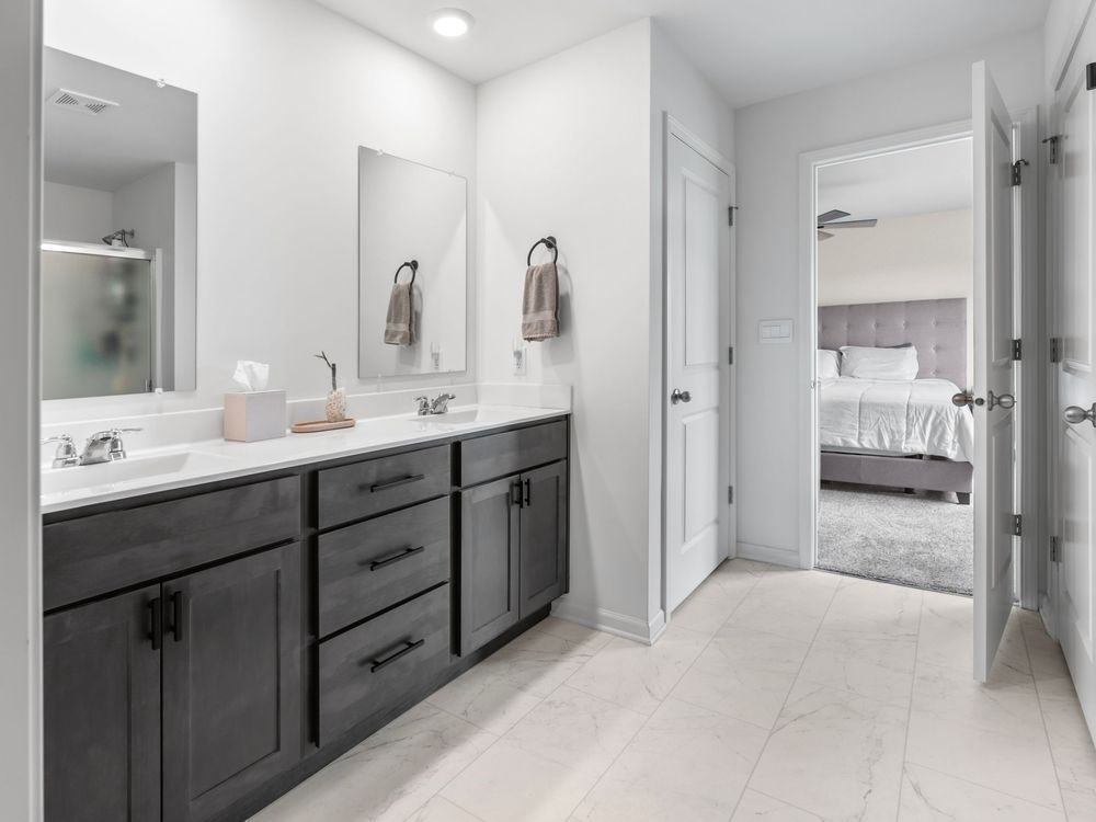 Modern bathroom with dark grey double vanity, white countertop, and light tile floors leading to a bedroom view.