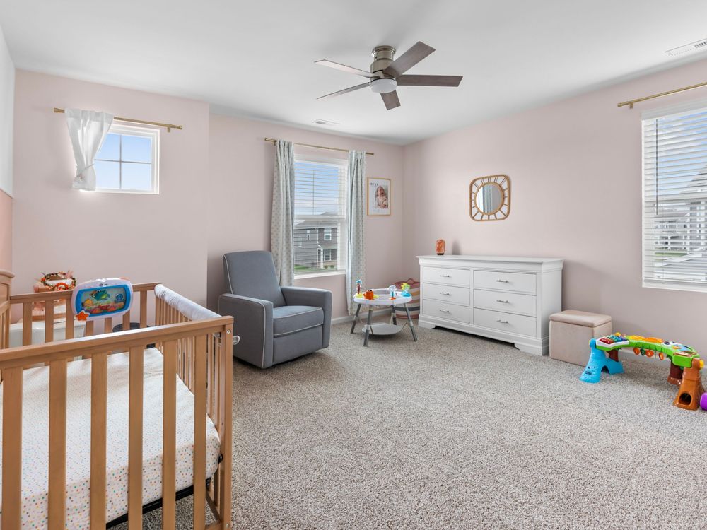 Light-filled nursery room with wooden crib, gray armchair, white dresser and carpeted floors.