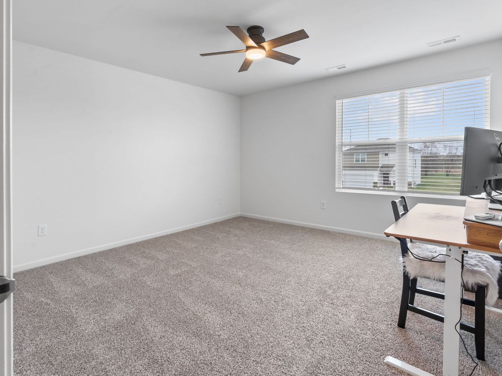 Empty bedroom or office space with beige carpet, white walls, window, ceiling fan, and small desk setup in corner.