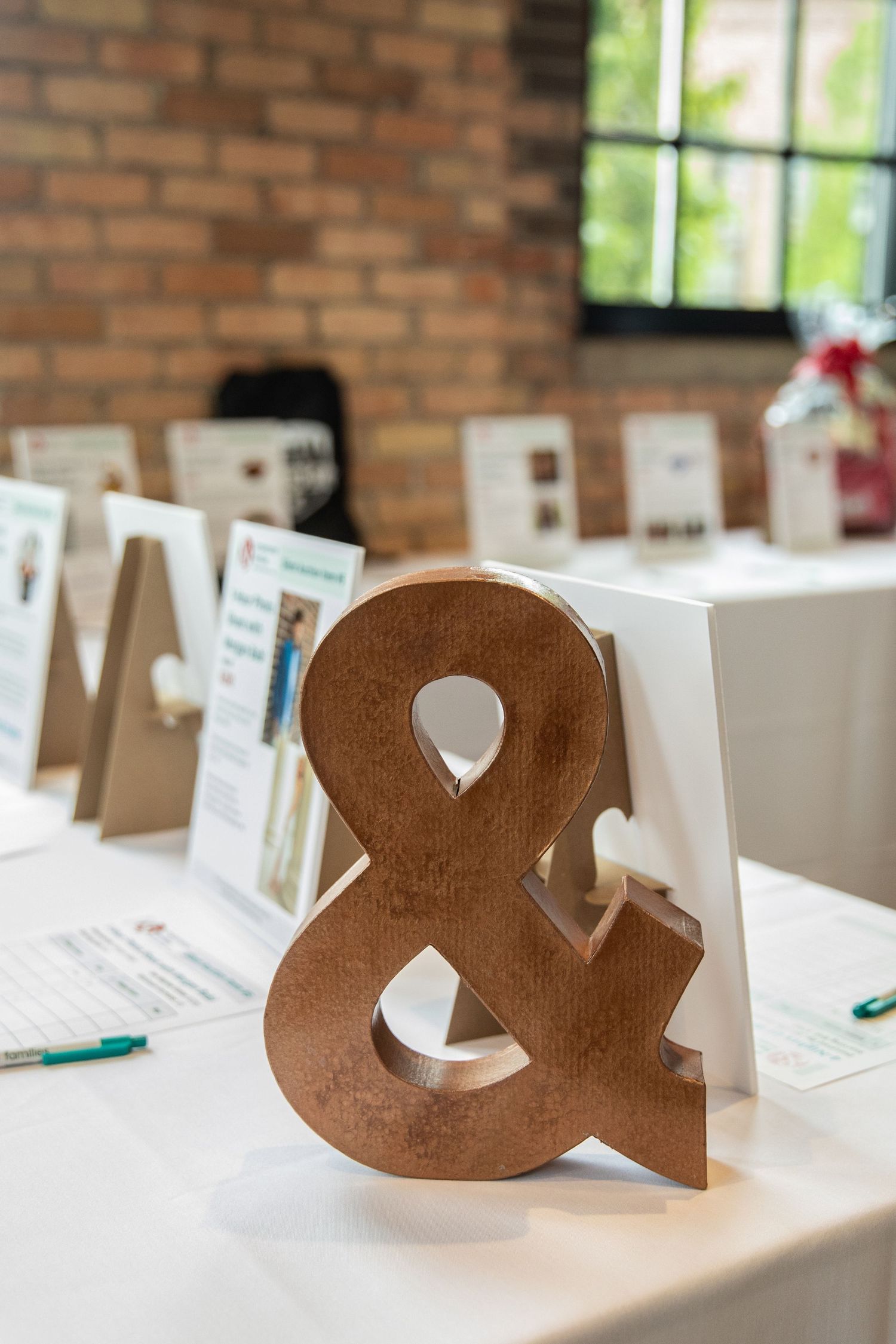 A wooden ampersand symbol sits on a white tablecloth at a wedding reception with place cards and decor in the background.