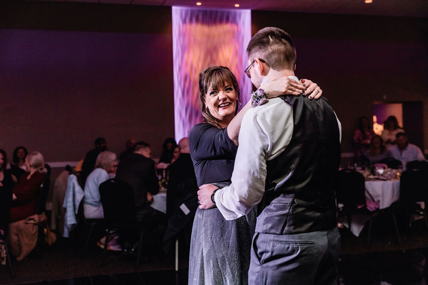 Dancing couple embrace and smile during a romantic moment at an evening reception with purple lighting and guests in background.