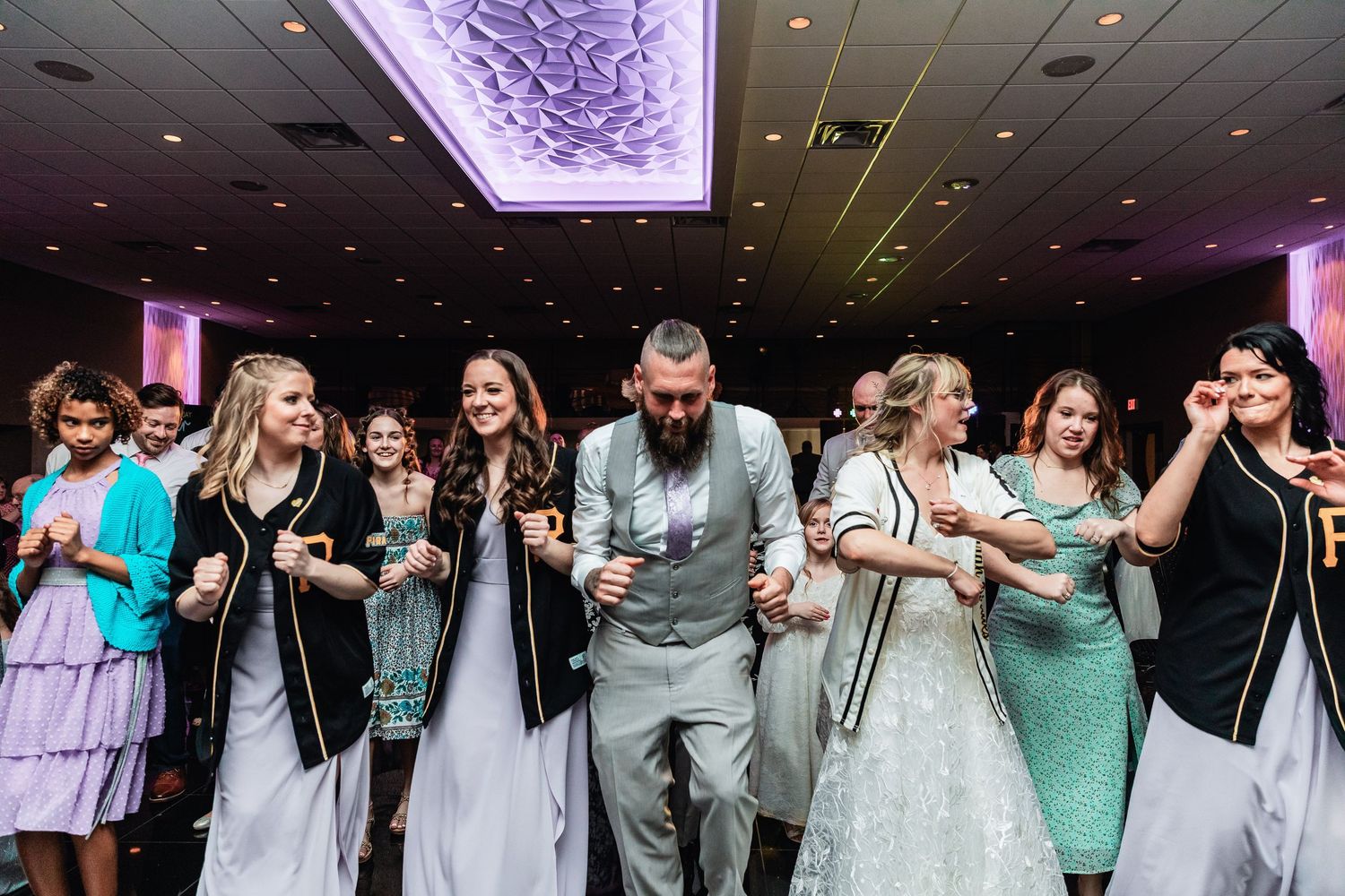 A group of wedding guests dance together under a purple-lit ceiling during a reception celebration.
