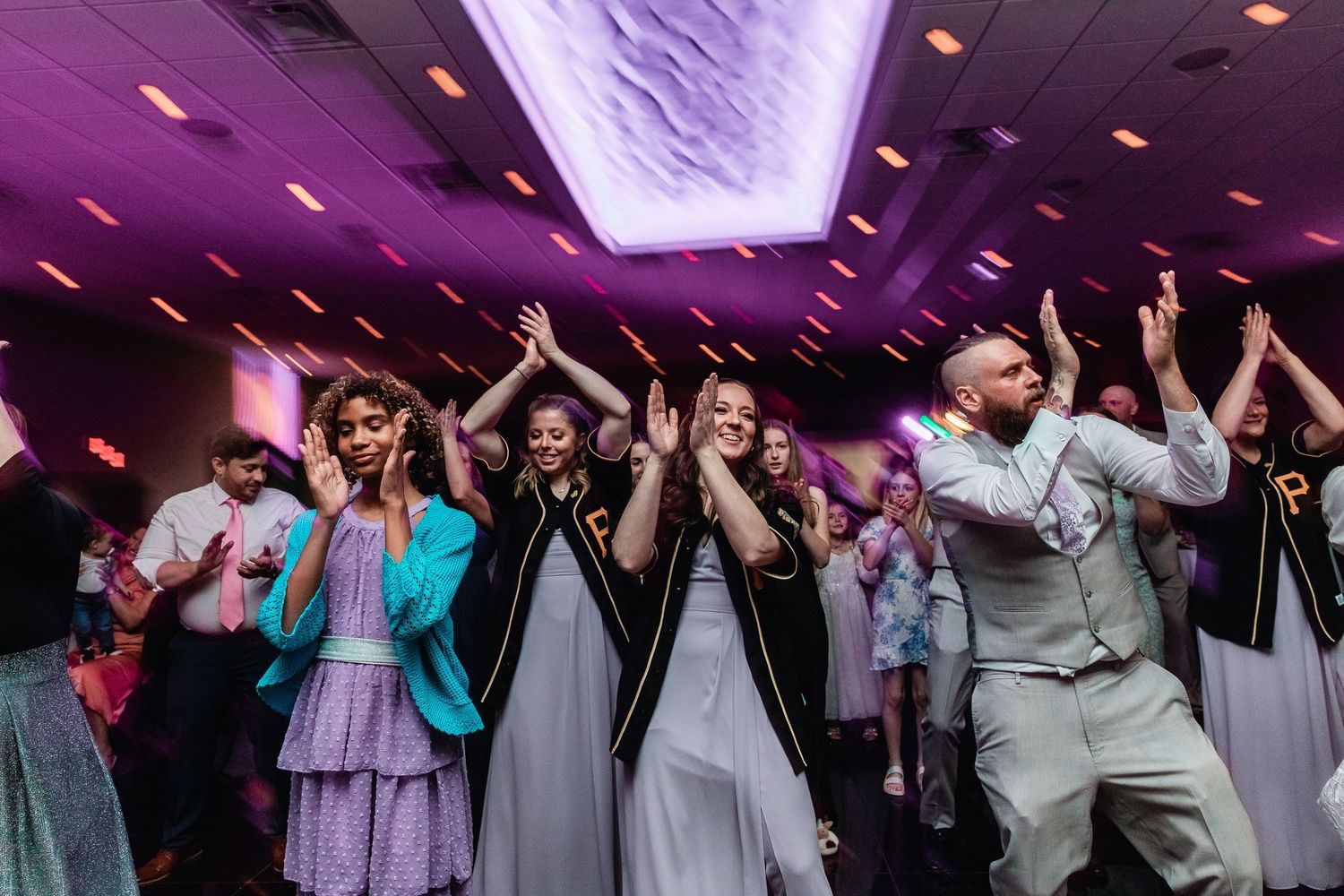 Wedding guests and attendees dance with hands raised during reception under purple lighting in celebration.