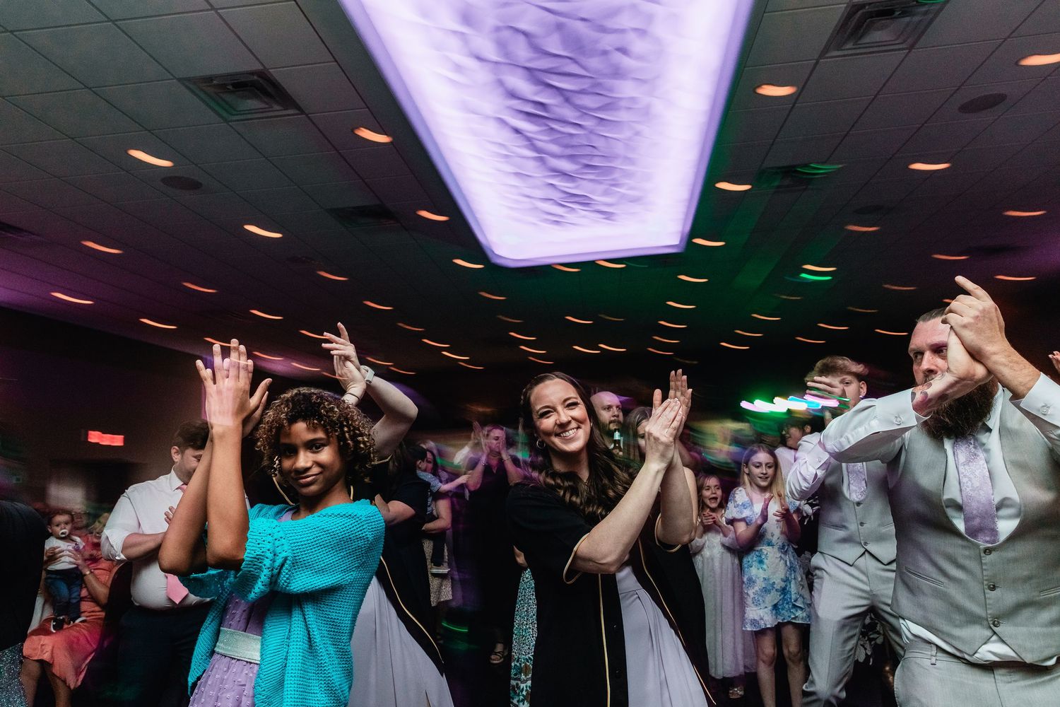 Wedding guests dancing enthusiastically at a reception under purple and green lighting on a dance floor.