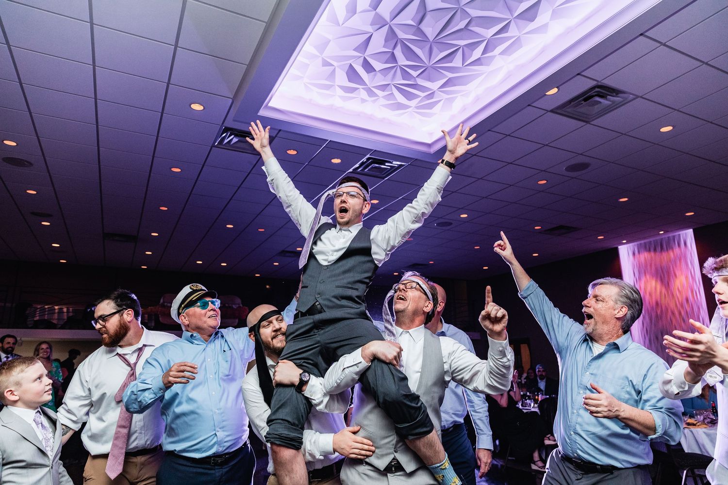 Energetic celebration in a ballroom with purple ceiling lights as someone jumps joyfully among cheering crowd.