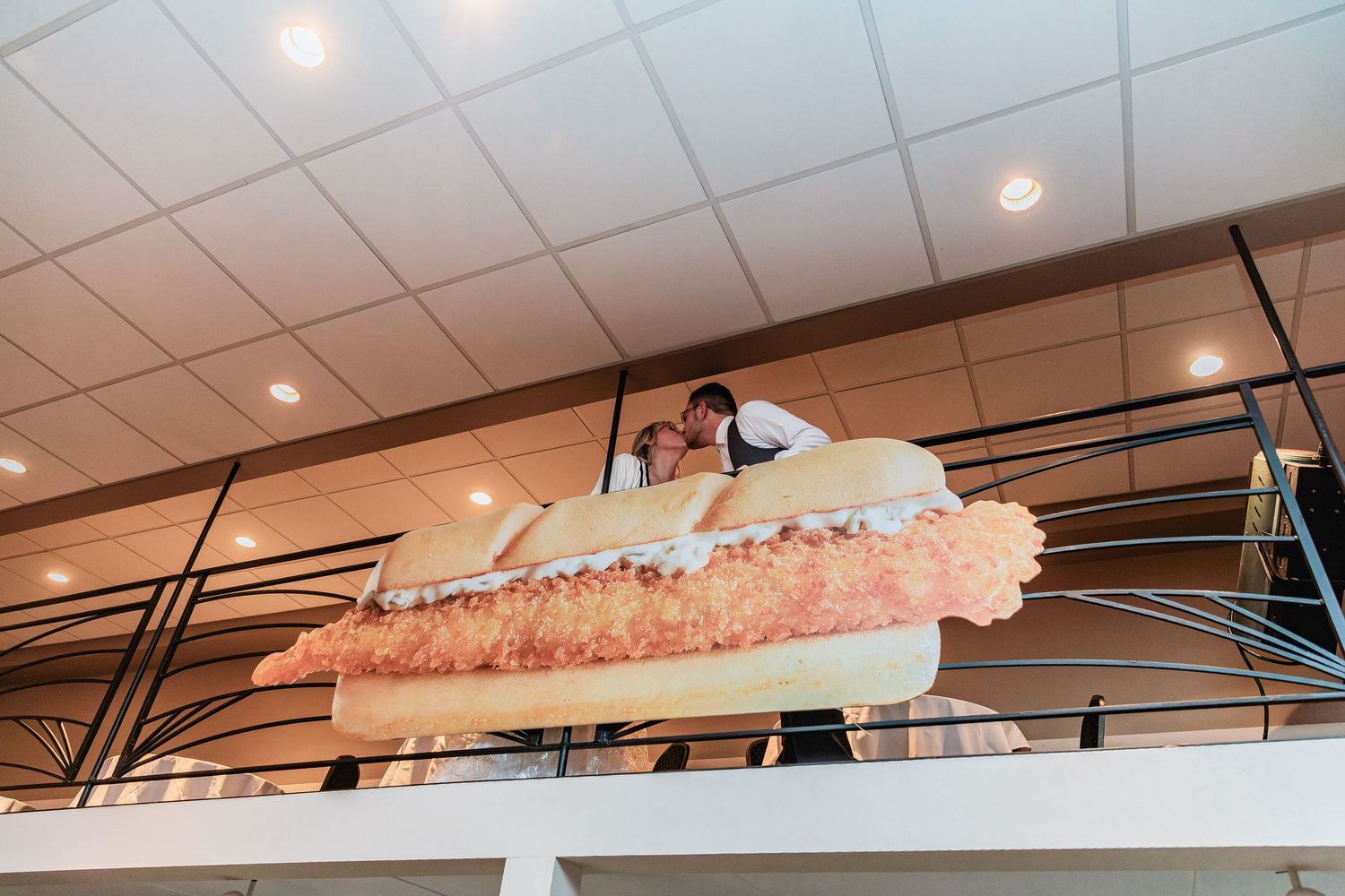 A couple poses atop a giant decorative pie sculpture mounted on an interior mezzanine wall with recessed lighting.