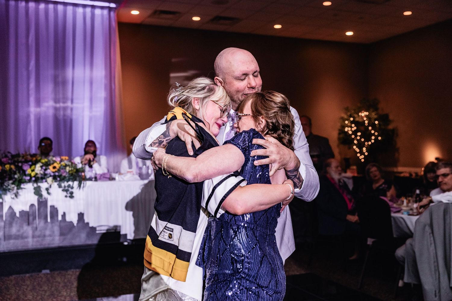 Emotional group hug on the dance floor at reception in elegantly decorated ballroom with purple lighting.