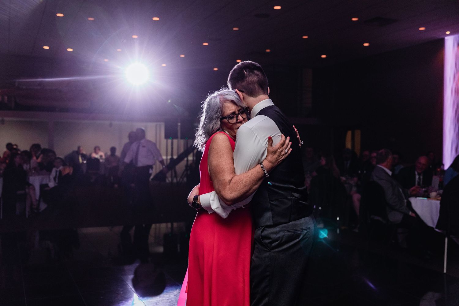 A couple shares an intimate dance in a dimly lit reception hall with dramatic backlighting creating a romantic silhouette.