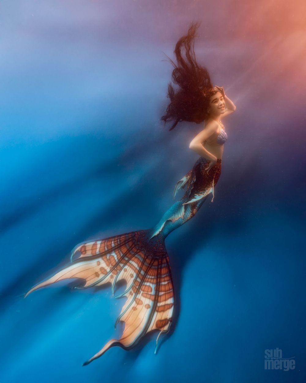 A mermaid with flowing hair swims gracefully through ethereal blue and orange underwater lights creating a dreamy atmosphere.
