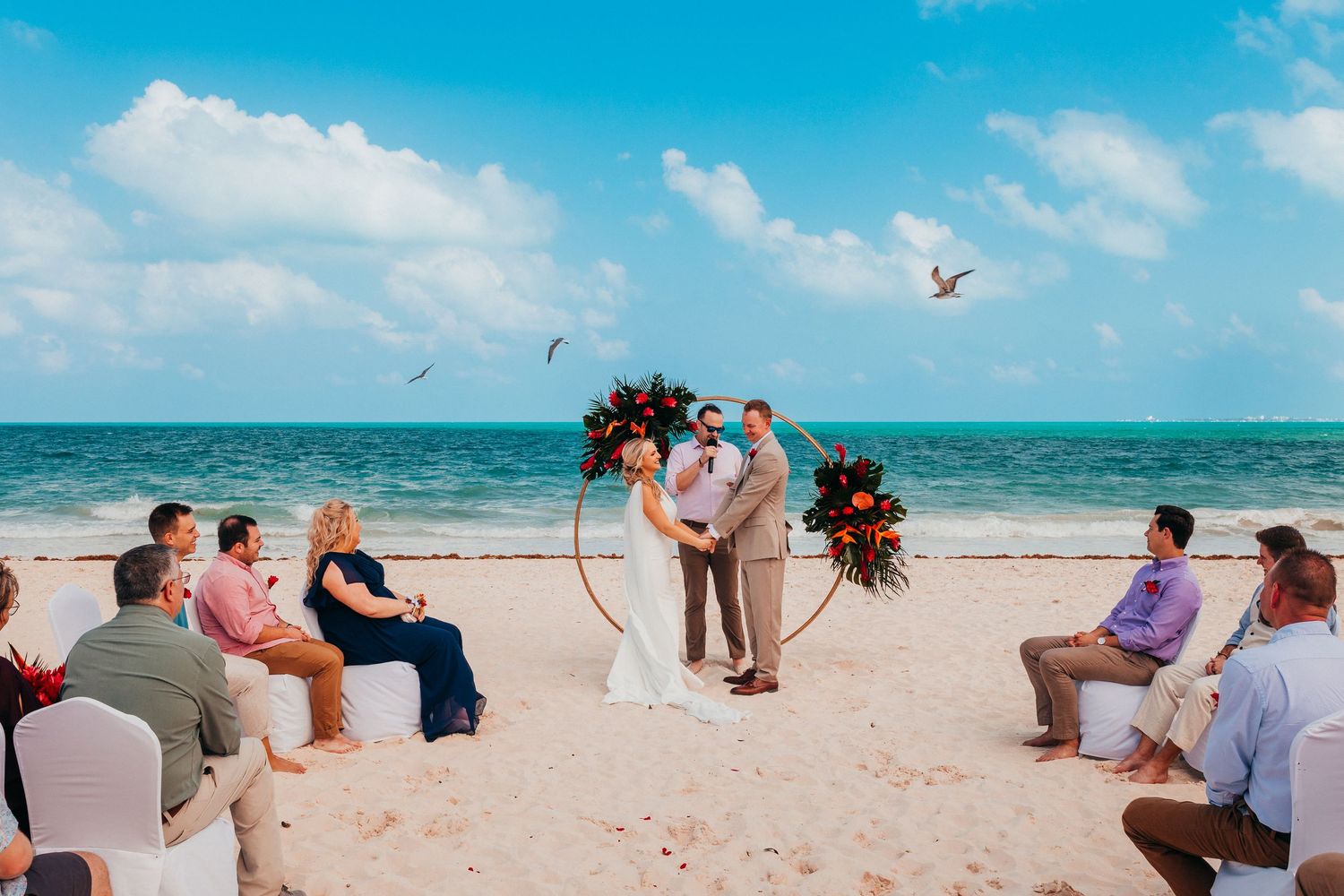 Small intimate beach wedding ceremony with guests seated on white chairs as couple exchanges vows by the ocean.