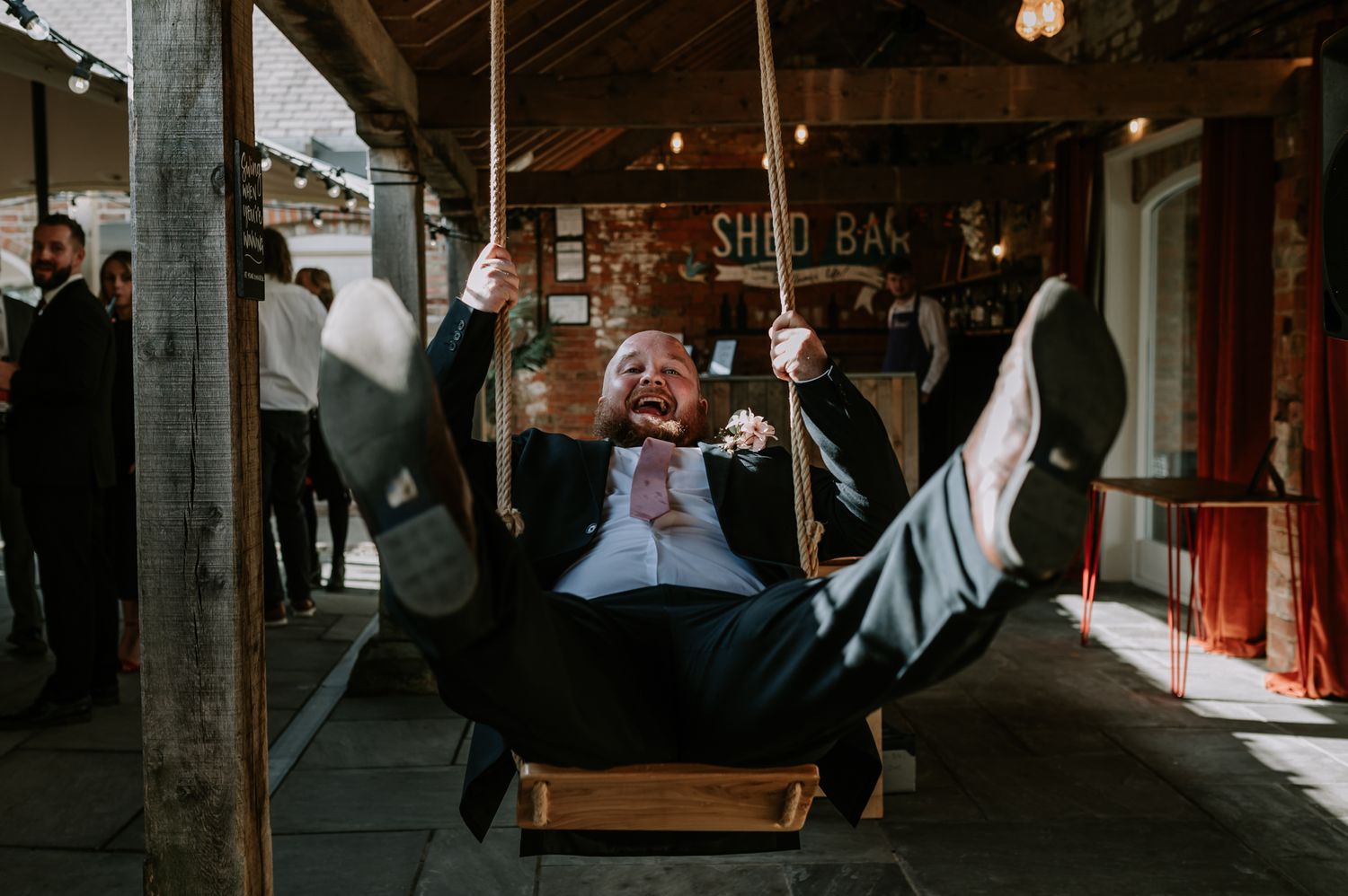 A man leans back on a swing with his legs spread, laughing and having fun at a wedding, looking directly at the camera.