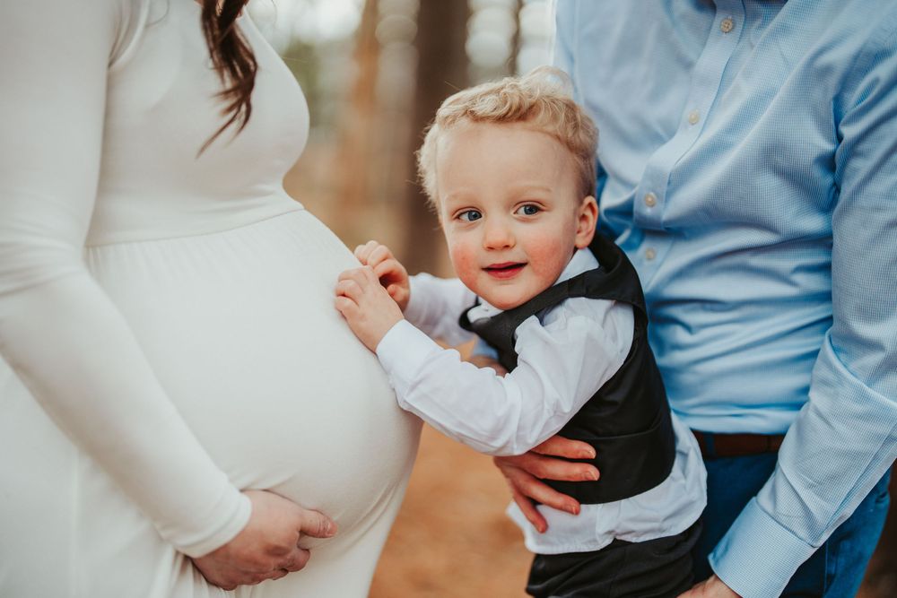 A young child in a bow tie and vest smiles while touching a pregnant woman's belly as someone looks on.